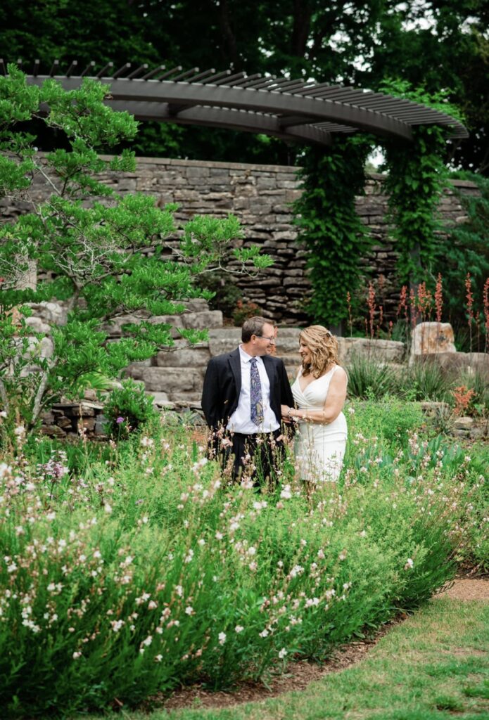 The bride and groom walk arm in arm in a garden full of lush greenery, pink flowers and potted plants. The bride is wearing a short  fitted white dress with a plunging neckline. The groom is wearing a black suit. They are at Cheekwood Botanical Garden in Nashville.