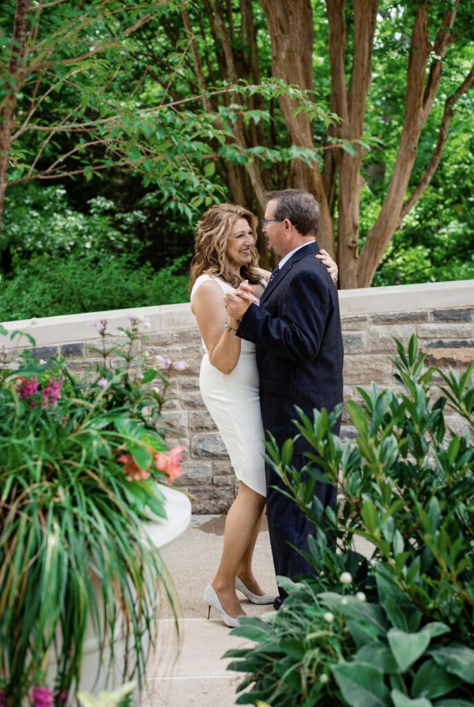 The bride and groom embrace as they dance in a garden full of lush greenery and pink flowers. The bride is wearing a fitted white dress with an asymmetrical hemline and plunging neckline. The groom is wearing a black suit with a white shirt and blue tie. They are at Cheekwood Botanical Garden in Nashville.