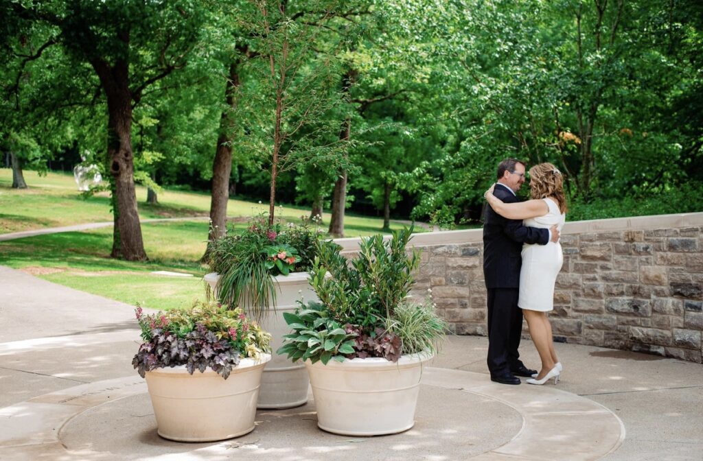 The bride and groom dance  in a garden full of lush greenery, pink flowers and potted plants. The bride is wearing a short  fitted white dress. The groom is wearing a black suit. They are at Cheekwood Botanical Garden in Nashville.