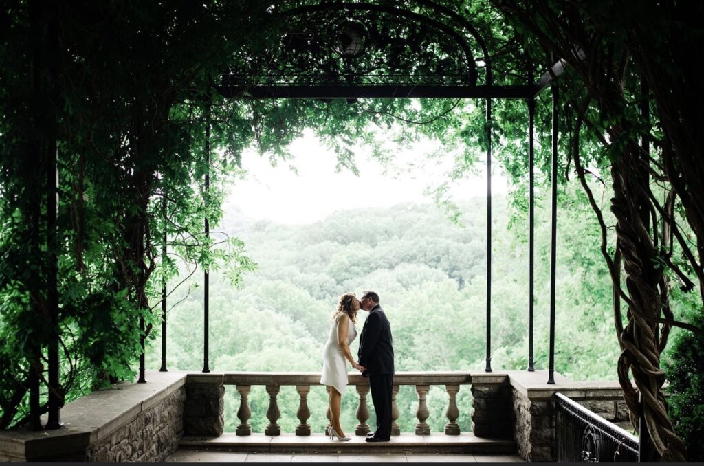 he bride and groom share a kiss in the Wisteria Arbor at Cheekwood Mansion. The bride is wearing a short fitted white dress with an asymmetrical hemline and a plunging neckline. The groom is wearing a black suit. The bride and groom are  holding hands as they lean in. They are framed by a large arbor of greenery and wisteria as they overlook lush green hills. They are standing on a stone balcony at Cheekwood Botanical Garden in Nashville.