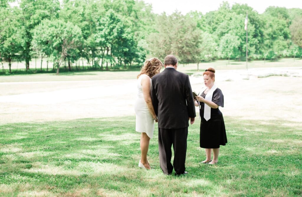 Groom wearing a black suit and the bride who is wearing a short, form fitting white dress with a plunging neckline face away from the camera as they listen to their officiant. Their wedding officiant is wearing a black dress with a white officiant sash and is reading from a book.