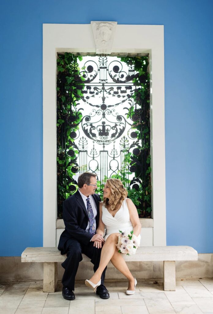 Sitting on a stone bench, the bride and groom smile up at each other. The bride is wearing a short fitted white dress with an asymmetrical hemline and a plunging neckline. The groom is wearing a black suit with a white shirt and blue tie. The bride is holding a bouquet of white, ivory and blush flowers. There is a black art deco decorative grate framed in the window behind them with bright blue walls on either side. They are at Cheekwood Botanical Garden in Nashville.
