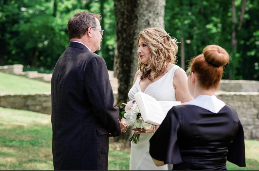 Groom wearing a black suit faces the bride who is wearing a short, form fitting white dress with a plunging neckline. The bride is holding a white bouquet. Their wedding officiant has her back to the camera. 
