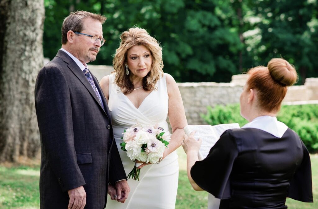 Groom wearing a black suit and the bride who is wearing a short, form fitting white dress with a plunging neckline face their officiant. The bride is holding a white bouquet. Their wedding officiant has her back to the camera. 