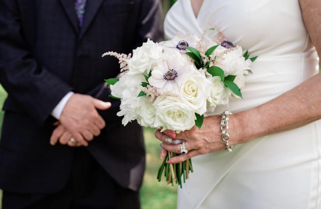 The bride shows off her white and blush bouquet. The bride is wearing a fitted white dress with a plunging neckline. Her nails are painted coral and silver. The bride is wearing a silver link bracelet, a diamond wedding band and a sapphire engagement ring. The groom is wearing a black suit.