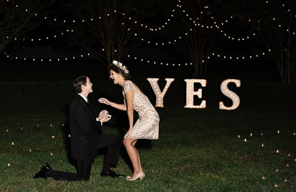 A man wearing a black suit kneels in the grass as he holds up an engagement ring. A woman in a short ivory and gold sequined dress with an ivory flower crown bends down towards him laughing. Large light up letters that say YES are in the background with string lighting hanging from the trees. 