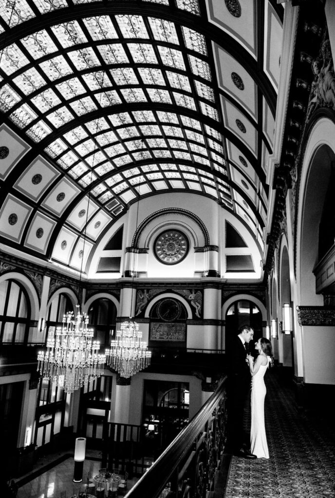 Black and white image of a bride in a form fitting floor length one shoulder sheath dress looking into the eyes of the groom who is wearing a black suit. They are standing on the balcony of the Union Station Hotel Nashville. There are two large crystal chandeliers and a barrel vaulted ceiling with stained glass. 