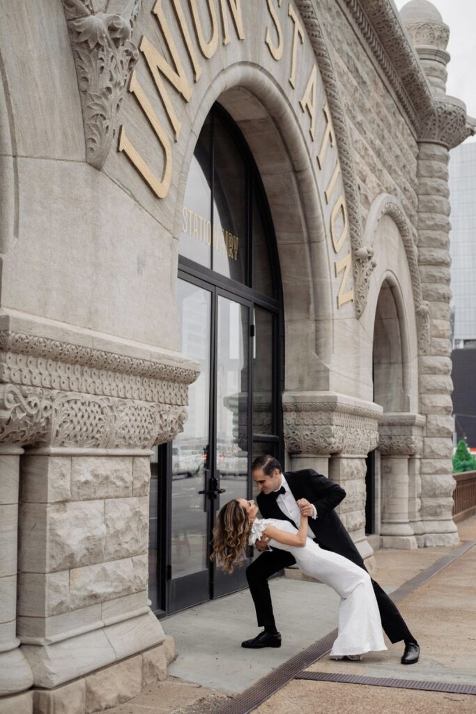 The bride in a form fitting floor length one shoulder sheath dress is dipped by the groom who is wearing a black tuxedo. He leans in to kiss her. They are standing in front of The Union Station Hotel. The rough stone of the building is to the left and the words Union Station are in gold above their heads. 