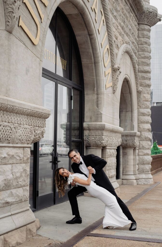 The bride in a form fitting floor length one shoulder sheath dress is dipped by the groom who is wearing a black tuxedo. They are both smiling at the camera. They are standing in front of The Union Station Hotel. The rough stone of the building is to the left and the words Union Station are in gold above their heads. 