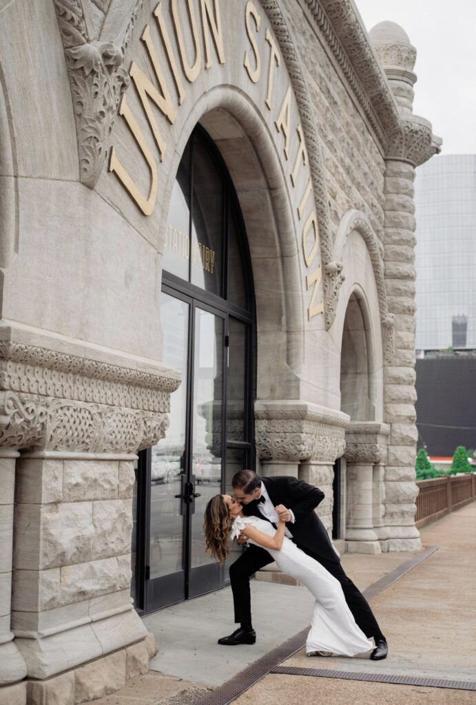 The bride in a form fitting floor length one shoulder sheath dress is dipped by the groom who is wearing a black tuxedo as he kisses her. They are standing in front of The Union Station Hotel. The rough stone of the building is to the left and the words Union Station are in gold above their heads. 