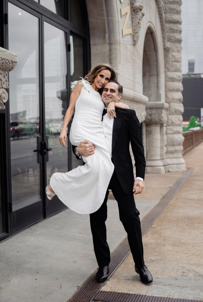 The bride in a form fitting floor length one shoulder sheath dress is being lifted up playfully by the groom who is wearing a black tuxedo. Her arm is wrapped around his neck and they are both smiling at the camera They are standing in front of The Union Station Hotel. The rough stone of the building is to the left. 