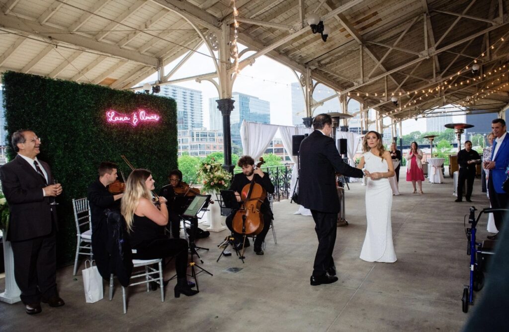 A string quartet called Viva La Strings Nashville plays music at the Union Station Hotel verandah. They are all dressed in black. They are sitting outside on a silver chiavari chairs as party guests dance to the music. 