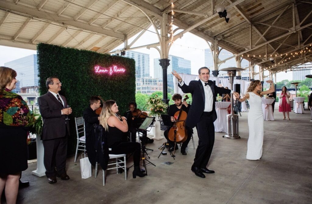 A string quartet called Viva La Strings Nashville plays music at the Union Station Hotel verandah. They are all dressed in black. They are sitting outside on a silver chiavari chairs as party guests dance to the music. A neon sign with Lana & Omar is lit up in pink behind the musicians. It hangs on a green boxwood wall.