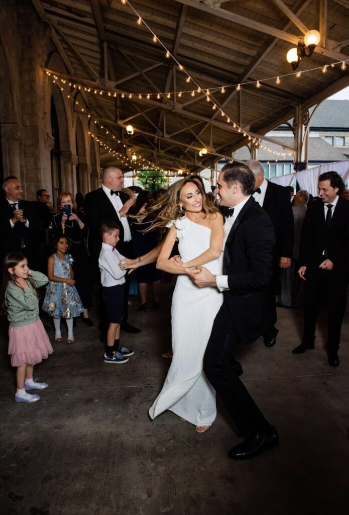 The groom spins the bride as they dance. The bride is wearing a floor length one-shoulder sheath dress. The groom is wearing a black tuxedo. String lights hang from the verandah ceiling above them. Guests crowd around to watch them.