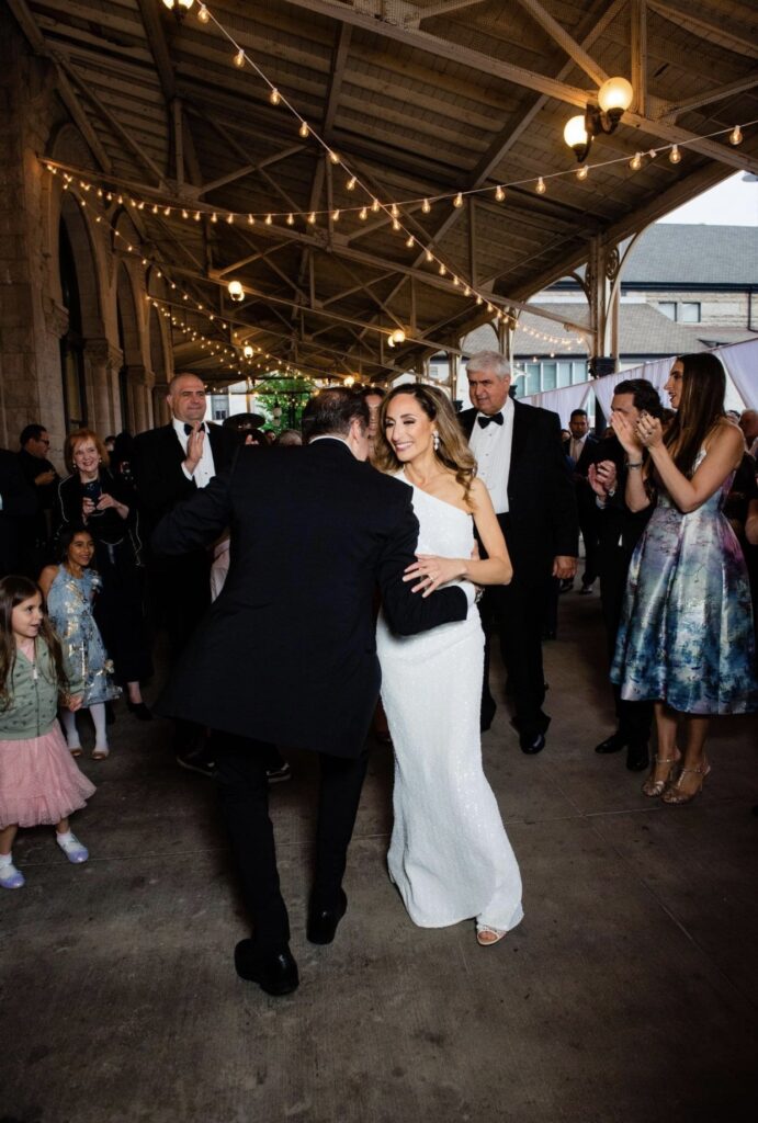 The groom leans in to kiss the bride as they dance. The bride, wearing a floor length one-shoulder sheath dress. The groom who is wearing a black tuxedo. String lights hang from the verandah ceiling above them. Guests crowd around to watch them.