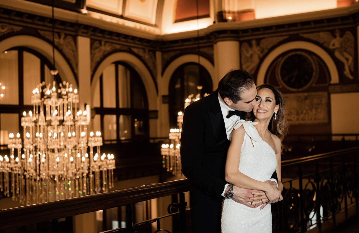 The bride in a form fitting floor length one shoulder sheath dress looks up at  the groom who is behind her wearing a black tuxedo. He kisses her on the cheek. They are standing on the balcony of The Union Station Hotel. There are two large crystal chandeliers behind them.  