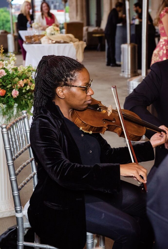 A viola player with Viva La Strings Nashville plays music at the Union Station Hotel. She is wearing a black jacket and black pants. She is sitting outside on a silver chiavari chair with a large floral centerpiece behind her. In the background party guests gather at cocktail tables to listen to the music. 