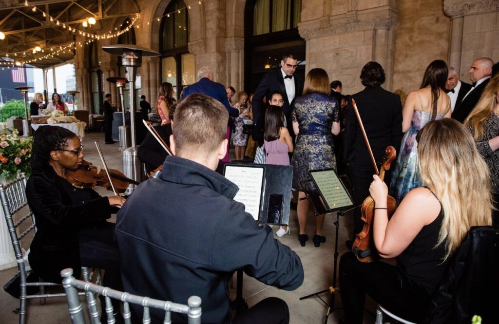 A violinist with Viva La Strings Nashville plays music at the Union Station Hotel. He is wearing a black shirt and black pants. He is sitting outside on a silver chiavari chair looking at his sheet music on an ipad. In the background party guests gather to listen to the music. 