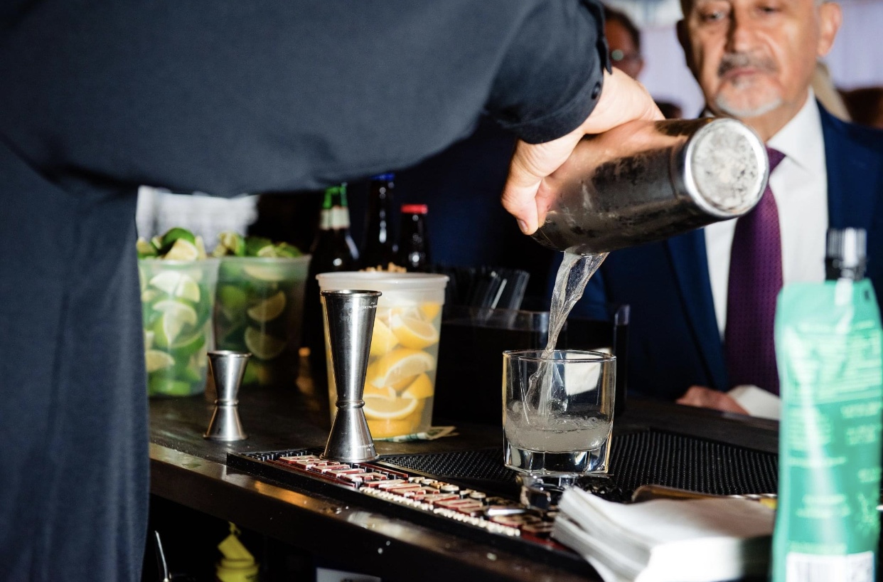 A bartender at the Union Station Hotel Nashville pours a cocktail from a silver shaker into a highball glass. There are cut up lemons and limes on the bar. The bartender is wearing a black uniform. A guest in a black jacket, white shirt and maroon tie waits for his drink.