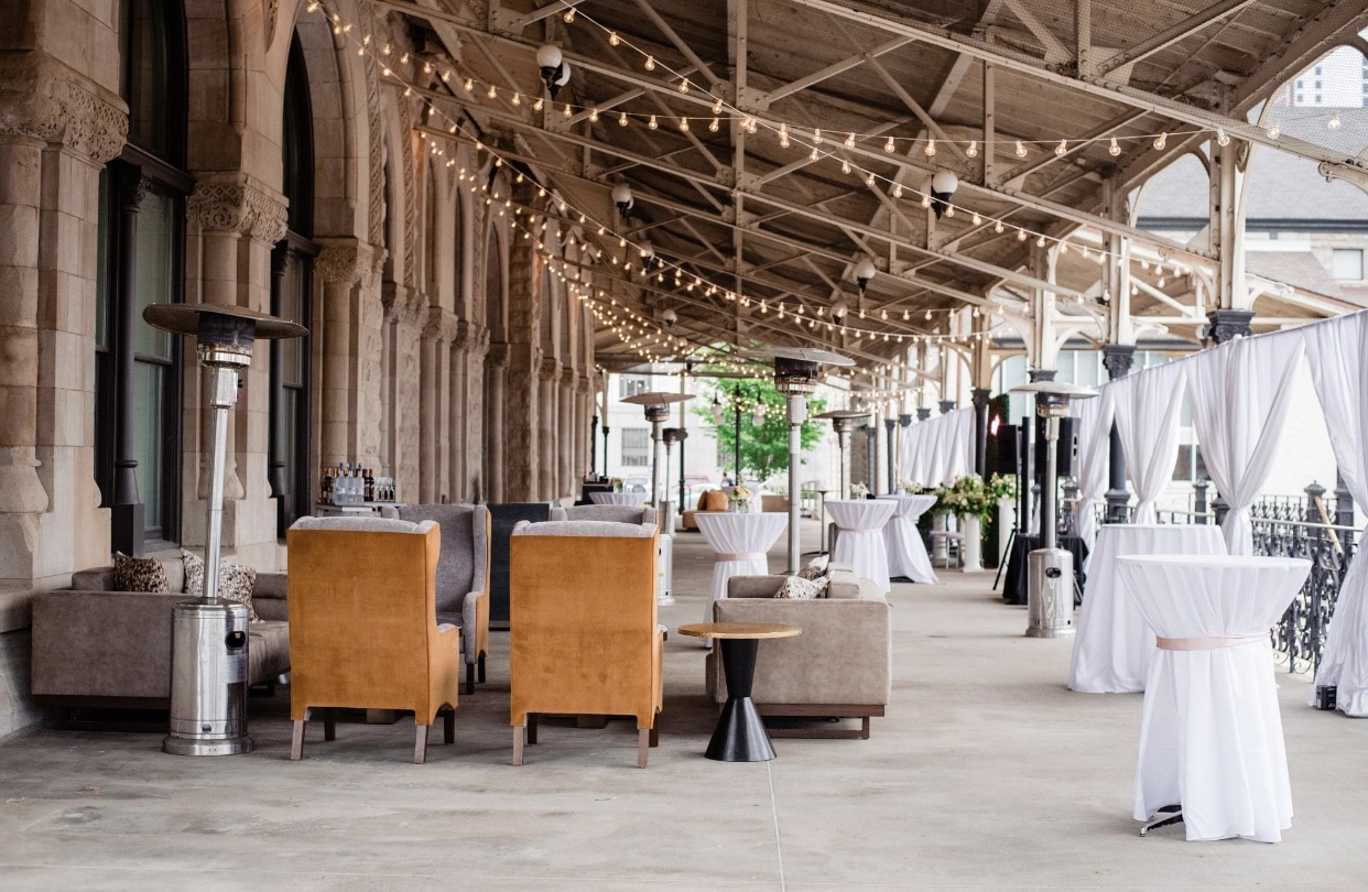 The verandah of the Union Station Hotel Nashville set up with rust colored arm chairs and soft khaki sofas accented by string lighting hanging from the ceiling. There is a  bronze and black coffee table off to the side and a tall silver umbrella heater. Cocktail tables with white linens and white drapery line the right hand side. 