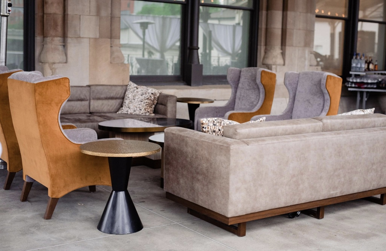 Gray and brown arm chairs and soft khaki sofas with bronze and white accent pillows set up on the verandah of the Union Station Hotel Nashville. There is a low wooden coffee table at the center of the soft seating area and a bronze and black coffee table off to the side. 