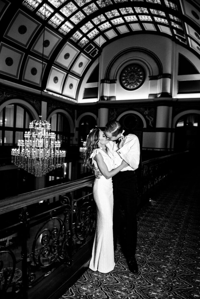 Black and white image of a bride in a form fitting floor length one shoulder sheath dress kissing the groom who is wearing a white shirt and black pants. They are kissing on the balcony of Union Station Hotel Nashville. There is a large crystal chandelier and a barrel vaulted ceiling with stained glass. 