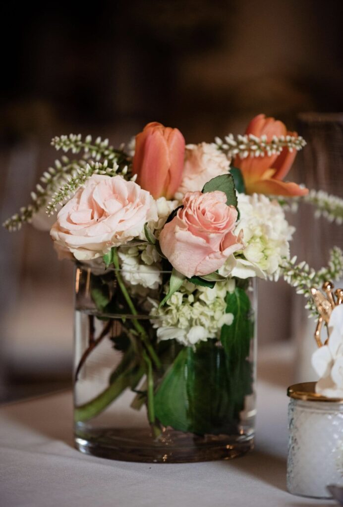 A small tabletop floral arrangement for a cocktail table in a glass cylinder sits on a white table cloth. The arrangement of flowers features pink roses, peach tulips, white hydrangea and veronica flowers.