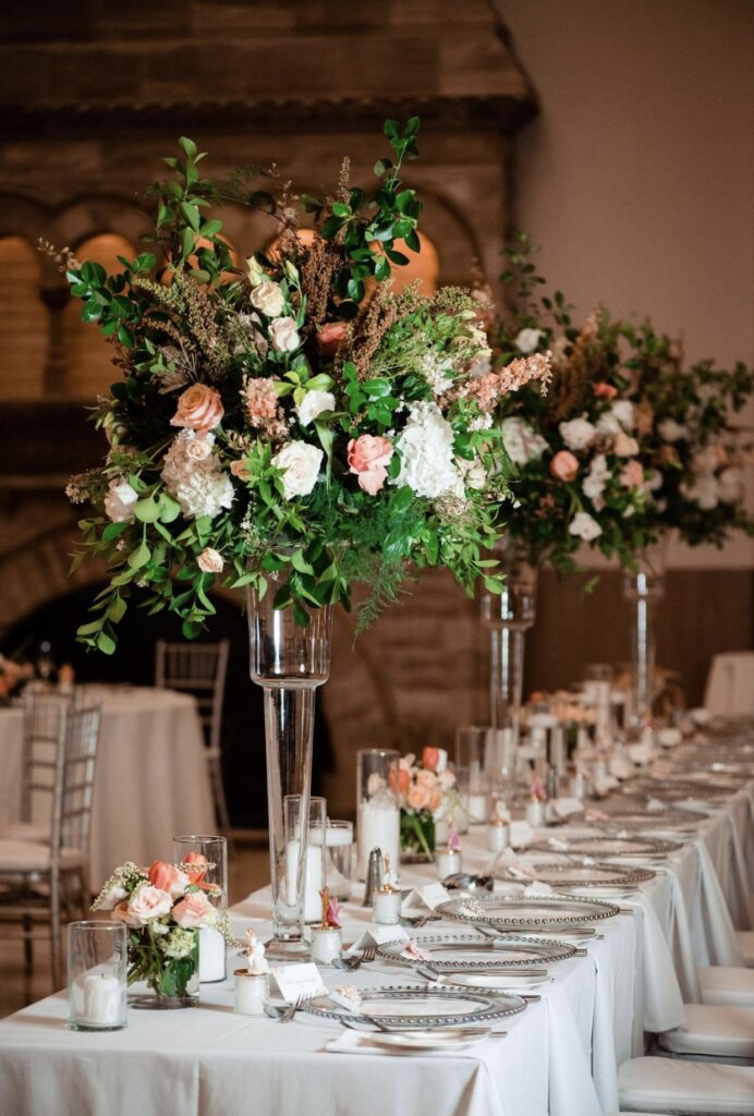 A long table with a white table cloth and clear glass chargers is set with white candles in glass cylinders and tall glass vases. Large floral arrangements of greenery, blush, pink and peach roses, tulips hydrangea, stock and ranunculus cascade down the sides. 