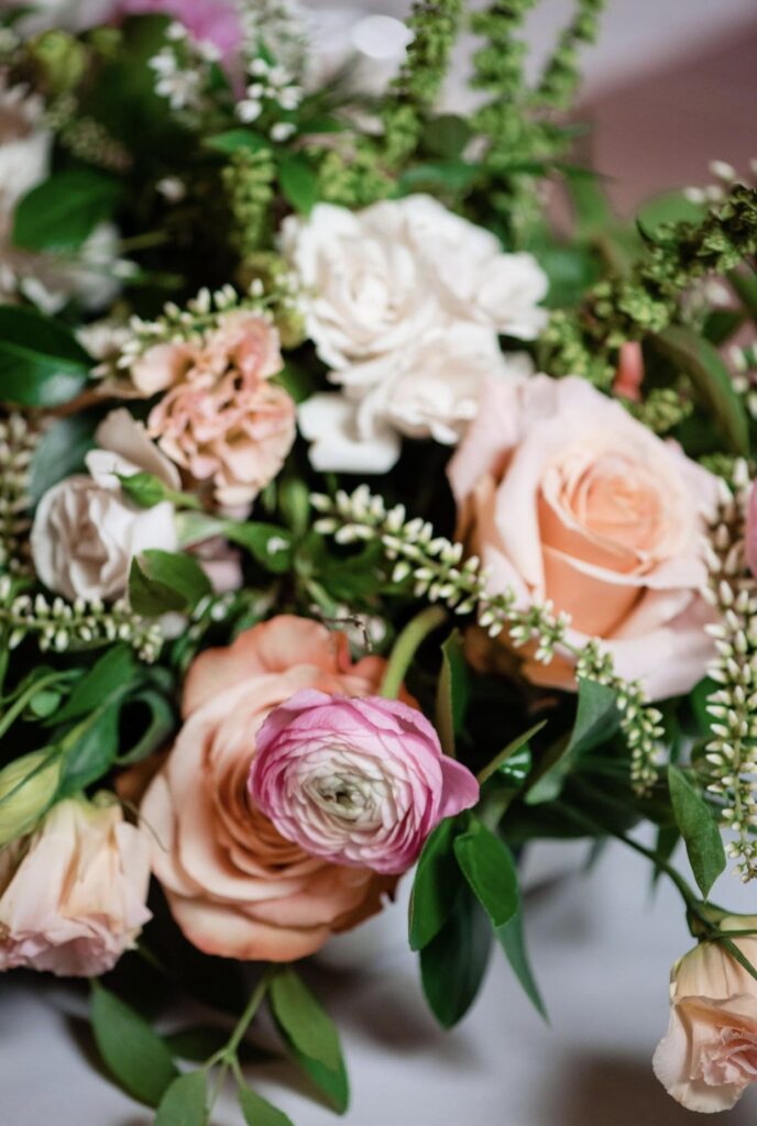 A detail image of a tabletop floral arrangement containing greenery, peach roses, hydrangea, veronica, and ranunculus.