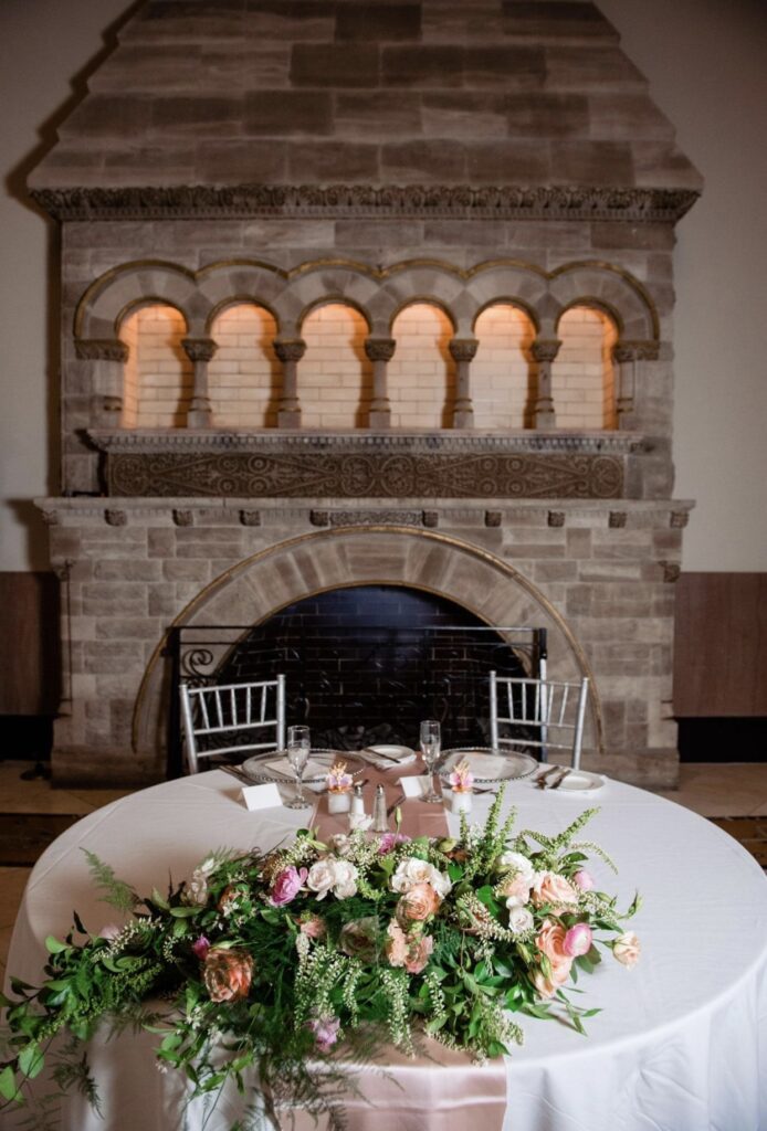 A round table with a white table cloth set for two guests with two silver chiavari chairs sits in front of a massive stone fireplace. On the table sits a low asymmetrical floral arrangement of greenery, blush, pink and peach roses, tulips hydrangea, stock and ranunculus.