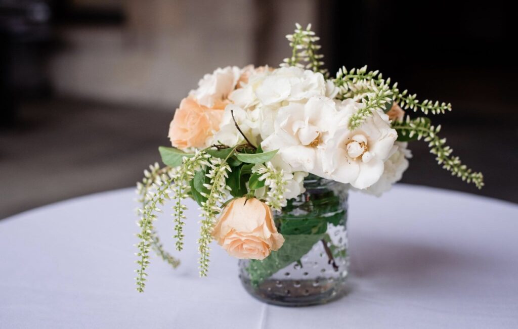 A small tabletop floral arrangement for a cocktail table in a beaded glass jar sits on a white table cloth. The bouquet features white roses, hydrangea, veronica, and peach tea roses.