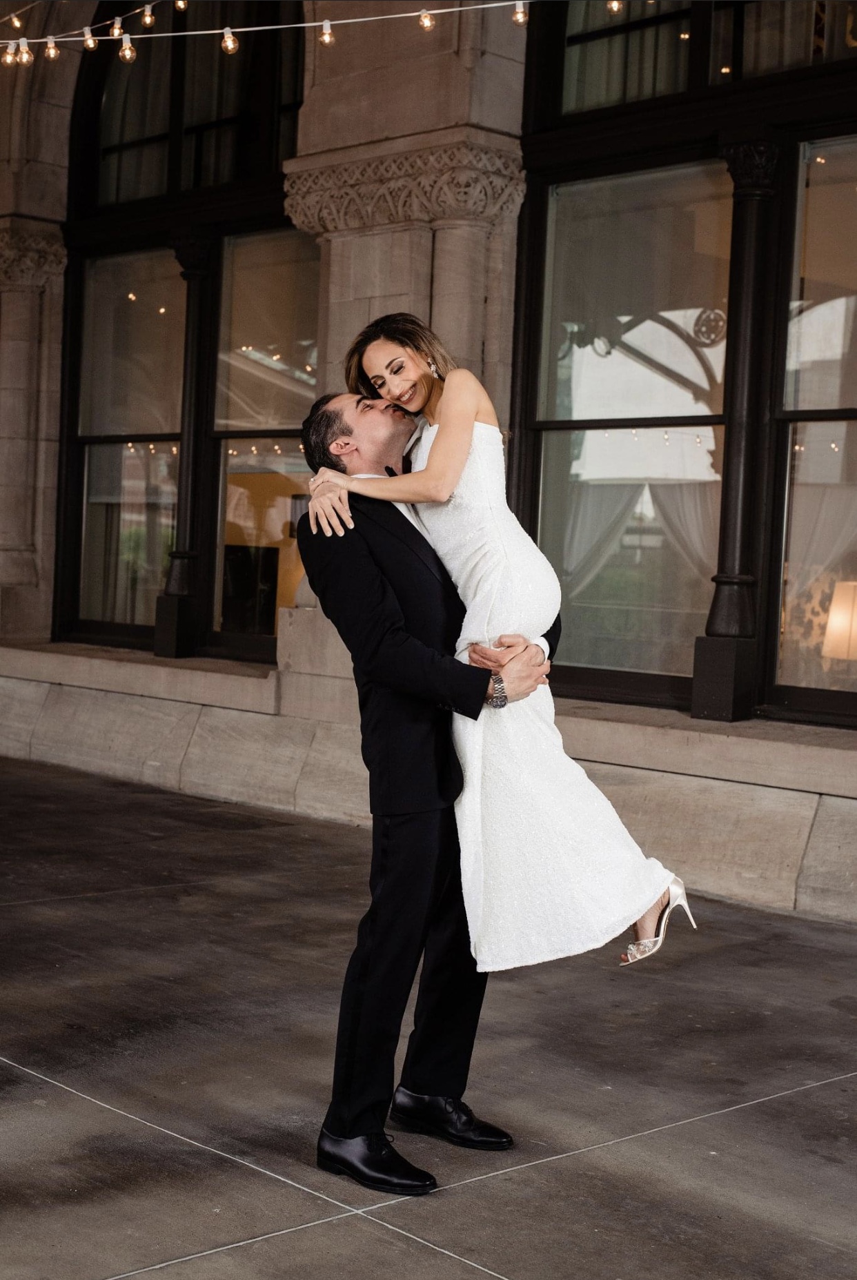 The bride in a form fitting floor length one shoulder sheath dress is lifted up by the groom who is wearing a black suit. He kisses her on the cheek. They are standing on the verandah of The Union Station Hotel. 