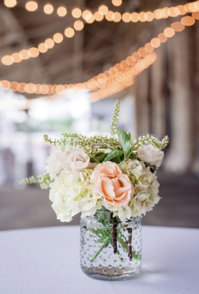 A small tabletop floral arrangement for a cocktail table in a beaded glass jar sits on a white table cloth. The bouquet features peach roses, hydrangea, veronica, and white tea roses. There are string lights hanging from the ceiling in the background.