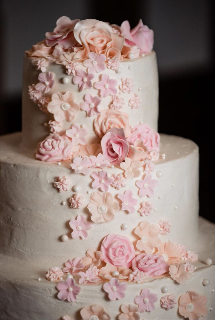 Detail of the upper half of a three tier wedding cake with ivory icing. The cake has peach, pink and blush sugar flowers cascading down the front. 