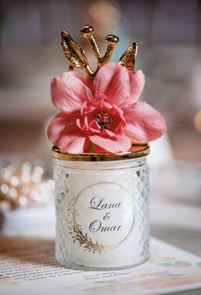 White votive candle in a cut crystal holder with a gold metal top. The top looks like a gold branch with a large pink silk flower. The words Lana & Omar are written on the front of the candle and are circled in gold with decorative leaves. 