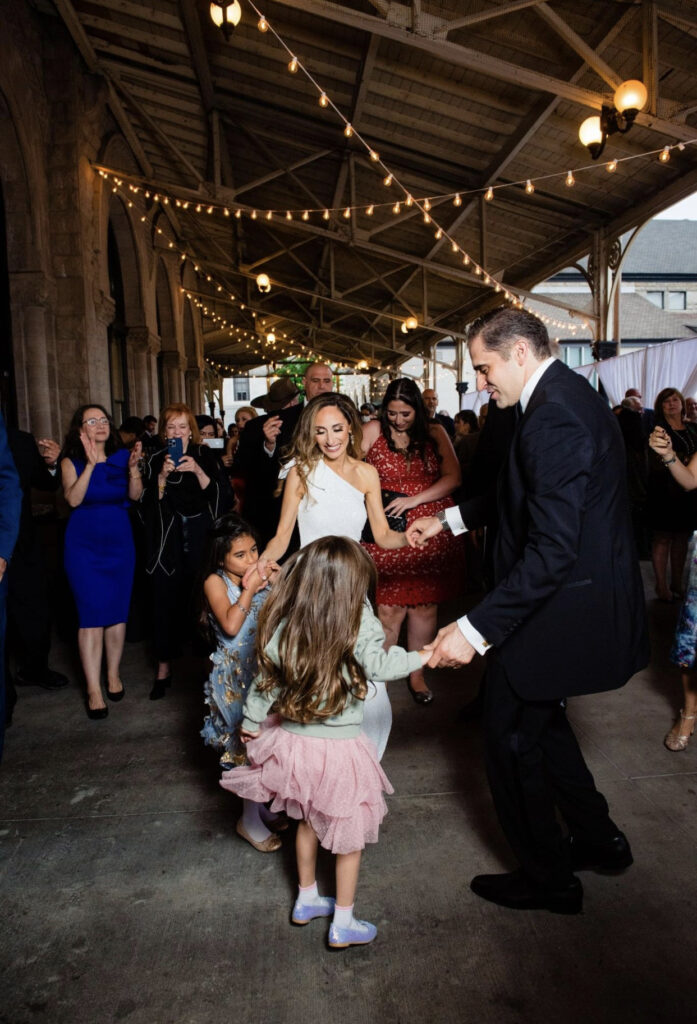 The bride, wearing a one shoulder dress and the groom wearing a black tuxedo dance with two little girls. They are at the Union Station Hotel Nashville on the verandah and there are string lights hanging from the ceiling. Other wedding guests watch the dancing in the background. 