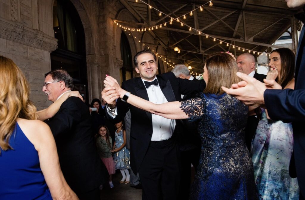 The groom, wearing a black tuxedo, dances with his mother. The mother of the groom is wearing a navy blue, short sleeved lace dress. Two little girls watch them dance in the background. They are at the Union Station Hotel Nashville on the verandah and there are string lights hanging from the ceiling and the stone arches are visible in the background. 