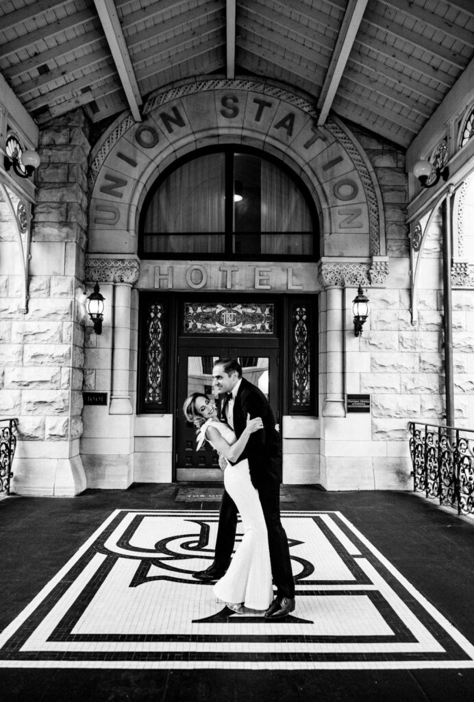 Black and white image of a bride in a form fitting floor length one shoulder sheath dress looking at the camera as the groom who is wearing a black suit looks at her. They are standing at the front entry of the Union Station Hotel Nashville. There is a mosaic floor with USH and a rough hewn stone wall with Union Station Hotel in an arch above their heads. 