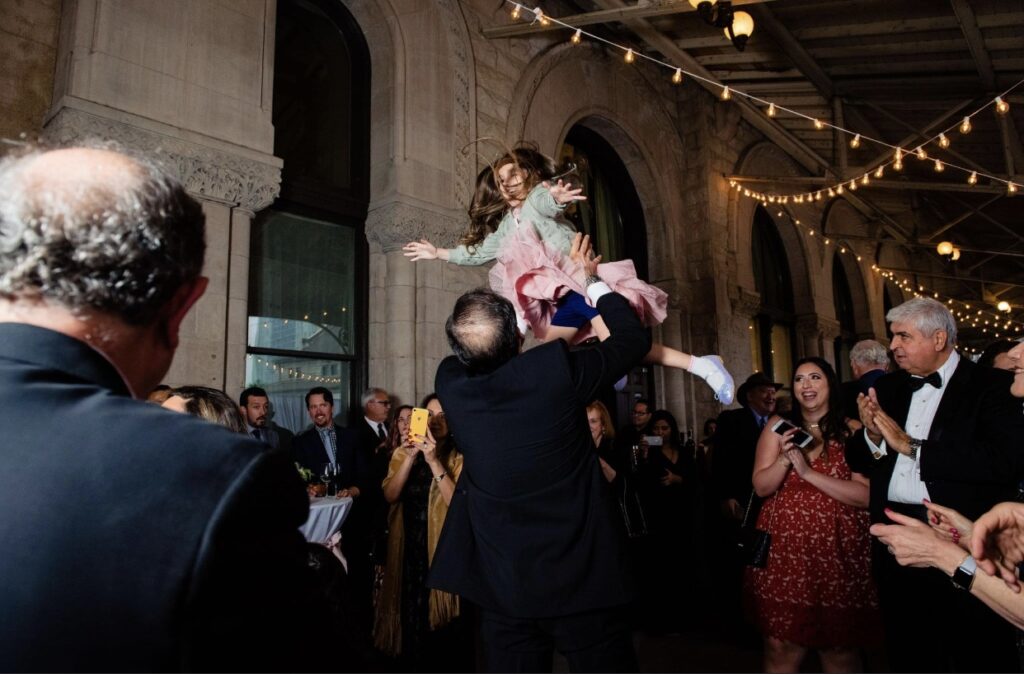 The groom wearing a black tuxedo tossed al little girl into the air as they dance. They are at the Union Station Hotel Nashville on the verandah. The stone arches are in the background and there are string lights hanging from the ceiling. Other wedding guests watch the festivities in the background. 