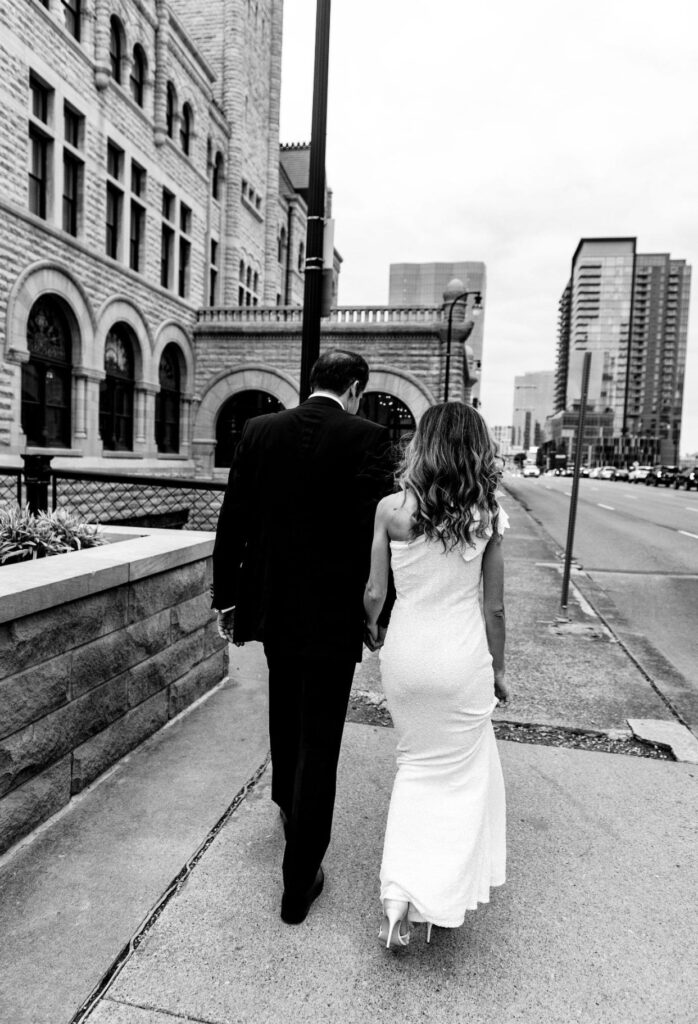 Black and white image of a bride in a form fitting floor length one shoulder sheath dress holding hands with the groom who is wearing a black suit  walk down the streets of Nashville. The Union Station Hotel is to the left. They are walking away from the camera. 