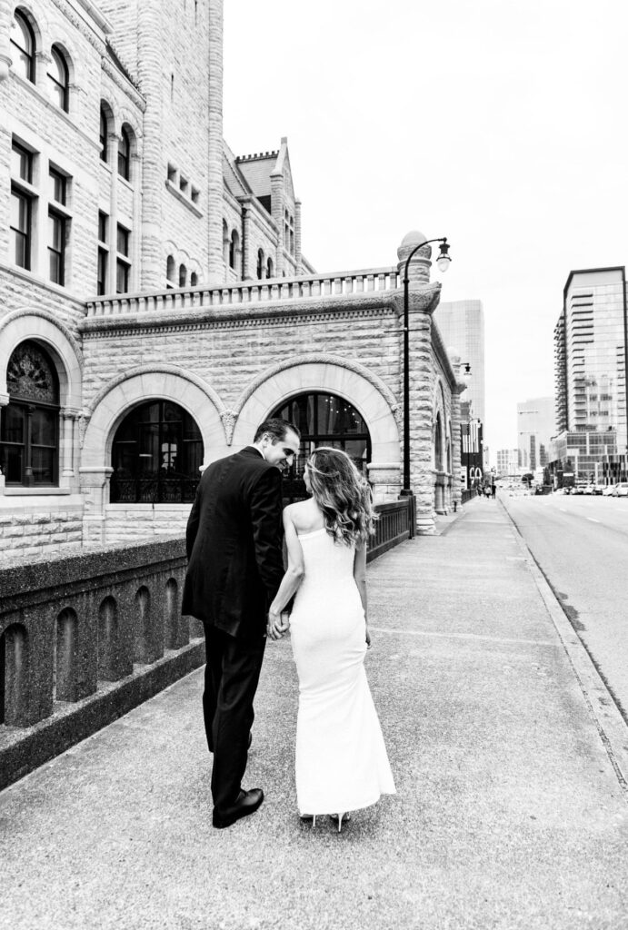 Black and white image of a bride in a form fitting floor length one shoulder sheath dress holding hands with the groom who is wearing a black suit  gaze into each other's eyes. The Union Station Hotel is to the left. They are facing away from the camera. 