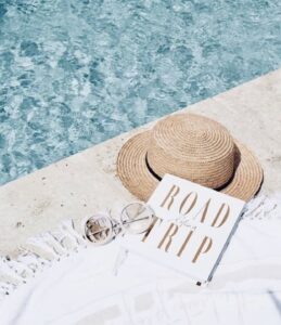 Road trip book on a beach blanket with white sunglasses and a straw sunhat next to the pool
