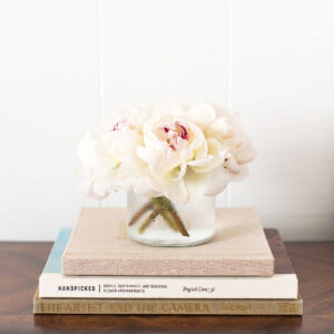 white peonies in a glass vase on top of a small stack of books