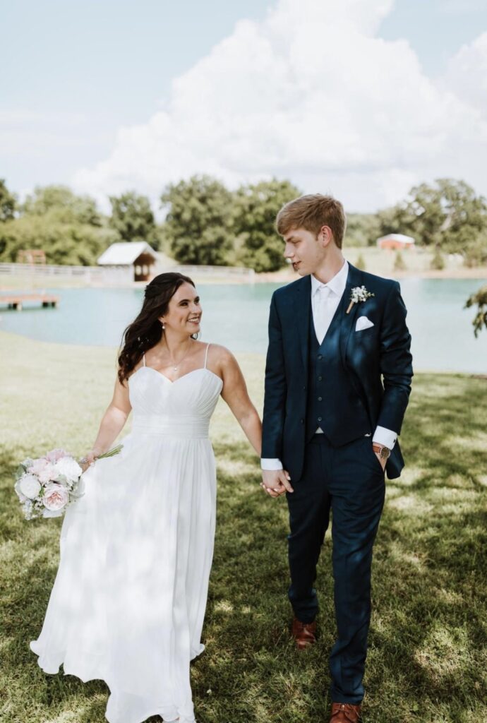 bride and groom walking beside a lake at Steel Magnolia Barn while holding a pink bouquet of flowers