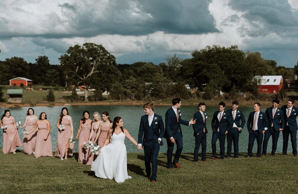 The bride and groom walk along the lakeside with their bridal party. The  groomsmen are dressed in navy suits and bridesmaids dressed in blush gowns holding pink and white bouquets.