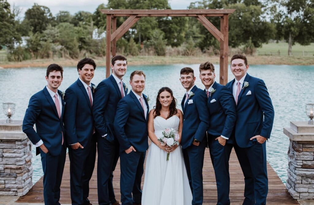 The bride stands on the lakeside dock with the groomsmen dressed in navy suits and blush ties and pocket squares. 