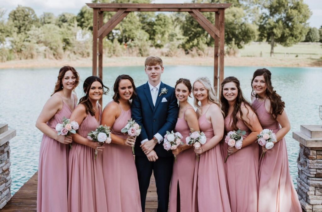 The groom stands on the lakeside dock with the bridesmaids dressed in blush and rose holding pink and white bouquets. 