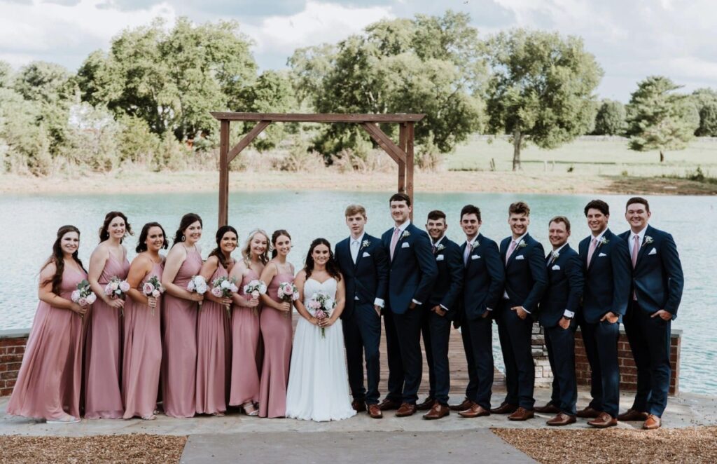 The bride and groom stand on the lakeside dock with the groomsmen dressed in navy suits and bridesmaids dressed in rose and blush gowns holding pink and white bouquets.