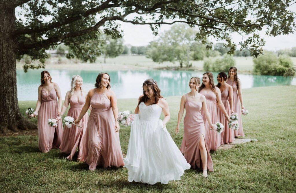 The bride walks along the lakeside with the bridesmaids who are dressed in blush gowns holding pink and white bouquets.