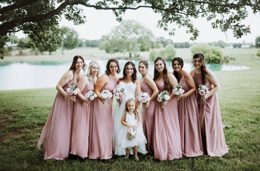 The bride poses along the lakeside with her bridesmaids and flower girl who are dressed in blush gowns holding pink and white bouquets.
